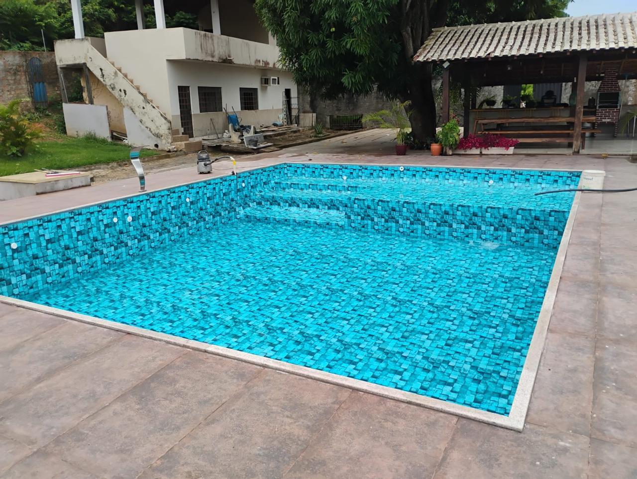 Piscina ampla de alvenaria revestida em pastilha azul-turquesa, com escada em degraus largos, cercada por piso de porcelanato e área de churrasqueira ao fundo.