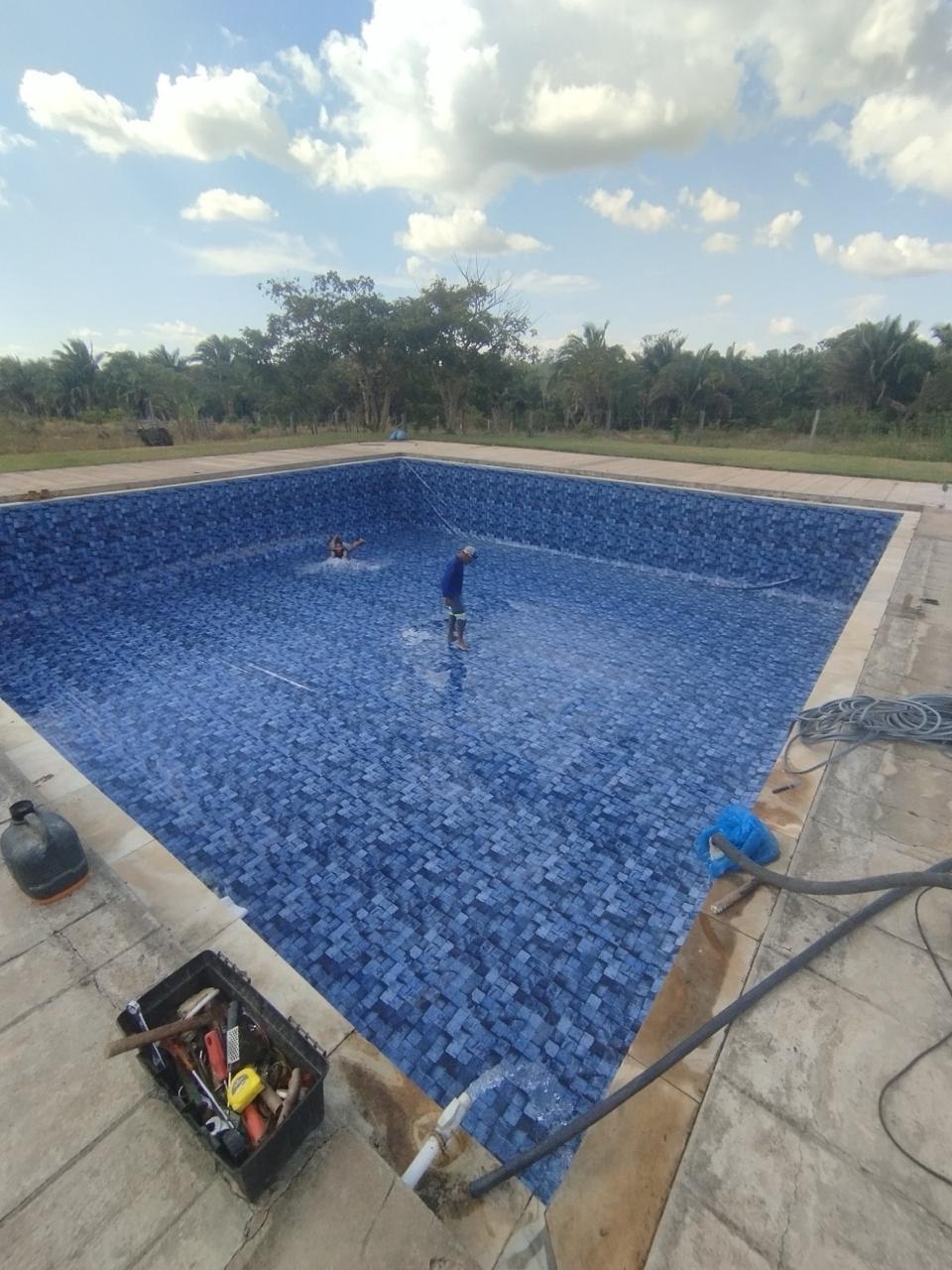 Piscina grande em pastilha azul-marinho durante o enchimento, com equipe finalizando a limpeza do fundo, em terreno aberto cercado de vegetação.