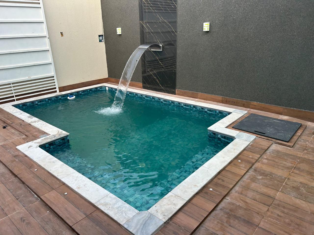 Piscina menor com água esverdeada e cascata de aço inox jorrando, cercada por deck de porcelanato amadeirado.
